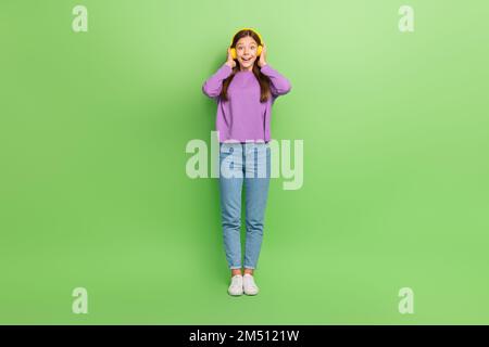 Langes Foto von süßen, schönen, positiv beeindrucken Mädchen tragen Denimjeans violette Pullover-Kopfhörer, die isoliert auf grünem Hintergrund tanzen Stockfoto