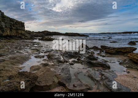 Eine felsige Bucht und ein Sandstrand unter einem ausdrucksstarken Himmel an der zerklüfteten Westküste Sardiniens Stockfoto