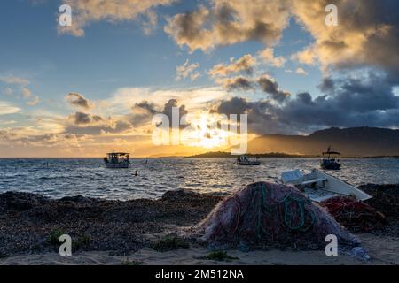 Pula, Italien - 10. Dezember 2022: Fischerboote vor Anker an der zerklüfteten Bergküste Sardiniens bei Sonnenuntergang mit Fischernetzen im Vordergrund Stockfoto