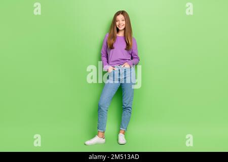 Großes Foto eines befriedigenden, bezaubernden Mädchens mit langem Haar, gekleidete Jeans, lila Pullover-Ärmel in Taschen, isoliert auf grünem Hintergrund Stockfoto