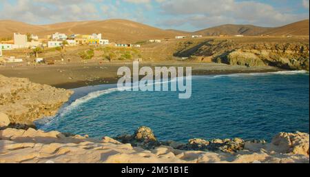 Spanien, Kanarische Inseln, Fuerteventura, vulkanischer Sandstrand in der Kleinstadt Ajuy. Blaue Ozeanwellen stürzen am schwarzen Meer ab. Stadtbild mit Resort Stockfoto