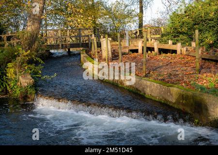 Wasserfall über ein kleines Wehr im Fluss mit einer alten Holzbrücke im Hintergrund Stockfoto