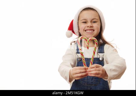 Ein liebenswertes kleines Mädchen, das den Weihnachtsmann trägt, mit roten und grünen Streifen Zuckerstangen in Herzform hält Stockfoto