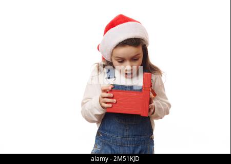 Es ist Zeit, die Weihnachtsgeschenke zu öffnen. Ein kleines Mädchen mit aufregendem Look packt die rote Geschenkbox mit einer glücklichen Überraschung aus Stockfoto