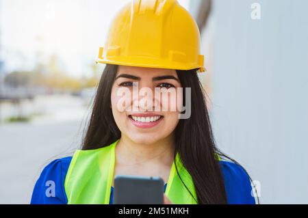 Vorderansicht Porträt einer jungen, weißhäutigen Ingenieurin mit gelbem Helm und reflektierender Weste, fröhliches Lächeln auf der Straße und Blick auf die Kamera, h Stockfoto