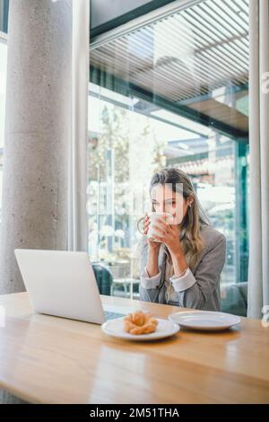 Junge, glückliche Geschäftsfrau, die Videoanrufe führt und Kaffee aus einer Tasse trinkt, in einem modernen, offenen Arbeitsplatz Stockfoto