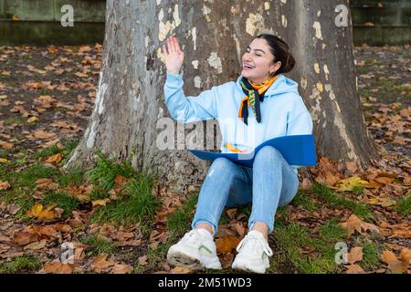 Ein orientalisches Mädchen sitzt unter einem Baum mit einem blauen Ordner in der Hand und lacht, Österreich Stockfoto