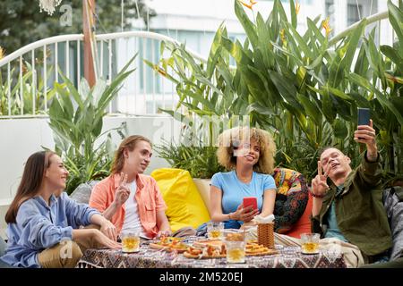 Fröhlicher junger Mann, der Selfie mit Freunden in einem Café im Freien macht Stockfoto