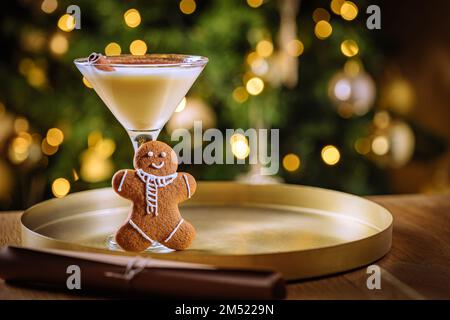 Eierpunsch-Cocktail in martini-Glas mit Lebkuchen-Mann. Getränk auf goldenem Tablett mit Weihnachtsbeleuchtung im Hintergrund. Silvesterfeier Stockfoto