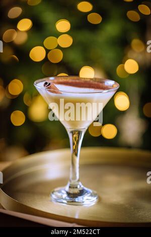 Eierpunsch-Cocktail in martini-Glas mit Lebkuchen-Mann. Getränk auf goldenem Tablett mit Weihnachtsbeleuchtung im Hintergrund. Silvesterfeier Stockfoto