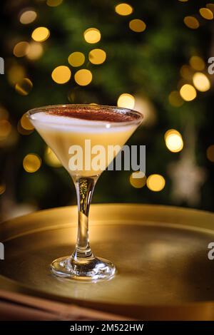 Eierpunsch-Cocktail in martini-Glas mit Lebkuchen-Mann. Getränk auf goldenem Tablett mit Weihnachtsbeleuchtung im Hintergrund. Silvesterfeier Stockfoto