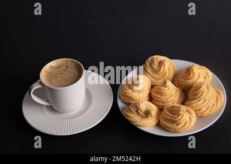 Fotografieren Sie Luftkuchen auf einem Teller neben Kaffee auf schwarzem Hintergrund Stockfoto