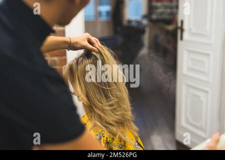 Der Stylingprozess mit Haarspray blonder Klientelhaare durch unerkennbaren, verschwommenen Friseur. Mittlere Nahaufnahme. Hochwertiges Foto Stockfoto