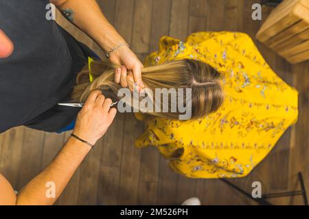 Draufsicht, wie ein Friseur die Haare seiner blonden Klientin kämmt, die einen gelben Umhang trägt. Hochwertiges Foto Stockfoto