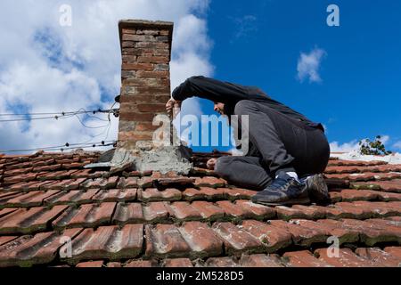 Ein erfahrener Handwerker auf dem Dach eines alten Hauses, der eine Kelle benutzt, um den Kamin zu reparieren. Der Mann sitzt konzentriert auf die anstehende Aufgabe. Himmel im Hintergrund Stockfoto