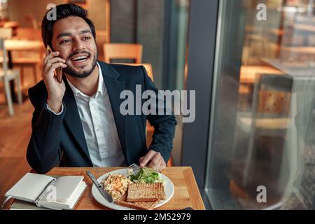 Ein gutaussehender indischer Geschäftsmann telefoniert während der Mittagspause in einem gemütlichen Café mit einem Kunden Stockfoto