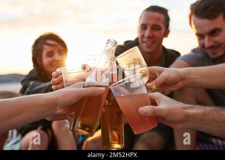 Auf den besten Sommer aller Zeiten. Eine Gruppe glücklicher junger Freunde, die mit ihren Drinks anstoßen, während sie sich am Strand entspannen. Stockfoto