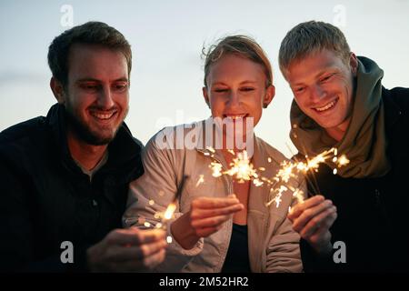 Freundschaft bringt den Funken zum Leben. Eine Gruppe glücklicher junger Freunde, die bei Sonnenuntergang am Strand mit Glitzern spielen. Stockfoto