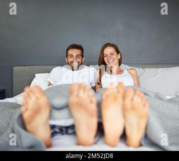 Oh, Sonntag, wie wir dich lieben. Ein glückliches, junges Paar, das sich im Bett entspannt, mit den Füßen, die unter der Bettwäsche herausstochen. Stockfoto