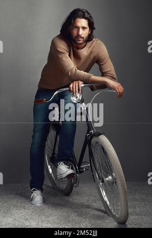 Meine bevorzugte Art der Reise. Studioaufnahme eines gutaussehenden jungen Mannes, der vor einem grauen Hintergrund gegen ein Fahrrad lehnt. Stockfoto