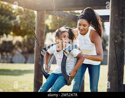 Der Park ist der Ort, an dem sie am meisten Spaß haben. Eine Mutter schiebt ihre Tochter auf eine Schaukel im Park. Stockfoto