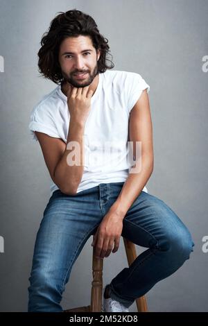 Nichts sieht besser aus als Selbstvertrauen. Studiofoto eines gutaussehenden jungen Mannes, der auf einem Hocker sitzt, vor grauem Hintergrund. Stockfoto