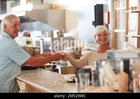 Es gibt einen Grund, warum ihre Kunden wiederkommen. Eine glückliche Seniorin, die einem Gast eine Tasse Kaffee in einem Café serviert. Stockfoto