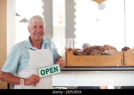 Ich bin bereit für dich. Porträt eines glücklichen Seniorenmannes, der ein offenes Schild in seiner Bäckerei hält. Stockfoto