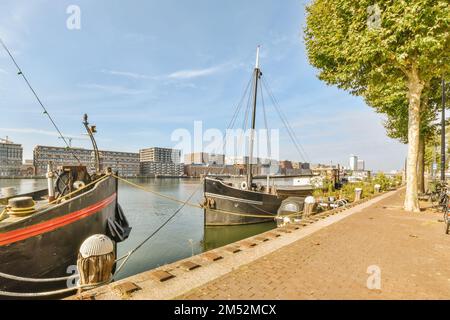 Zwei Boote, die im Wasser anlegen, mit Gebäuden im Hintergrund und Menschen, die auf dem Bürgersteig neben anderen Booten laufen Stockfoto