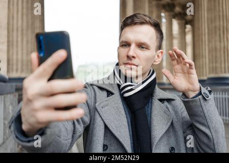 Der attraktive junge Mann im Mantel, der auf der Straße unterwegs ist, hält sein Handy und macht Videos oder Fotos, macht ein Selfie auf dem Smartphone und kommuniziert online über Videokommunikation Stockfoto