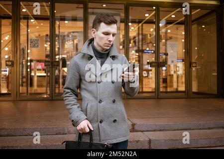 Ein lächelnder junger Mann in einem grauen Mantel hält Scroll-SMS auf seinem Handy auf der Straße im Stadtzentrum. Ein lächelnder Mann verlangt ein Taxi in der Anwendung. Stockfoto