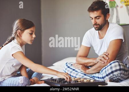 Dad hat ihr gut beigebracht, Dame zu spielen. Ein junger Vater, der ein Brettspiel mit seiner süßen kleinen Tochter zu Hause spielt. Stockfoto