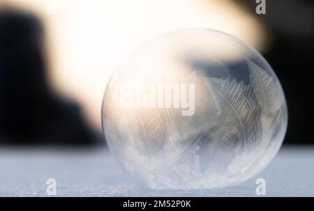 Nahaufnahme einer gefrorenen Seifenblase. Der Ball liegt auf Schnee. Der Hintergrund ist dunkel, und dazwischen helles Licht. Die Kuppel ist durchsichtig Stockfoto