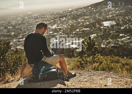 Gehen Sie dorthin, wo Inspiration Sie hinführt. Ein junger Mann, der seinen Laptop benutzt, während er auf einem Berggipfel sitzt. Stockfoto