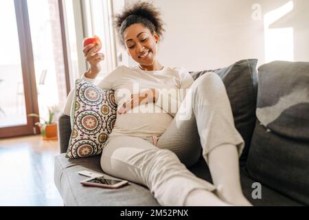 Erwachsene, wunderschöne, lächelnde afrikanische Frau, die Apfel isst und ihren Bauch hält, während sie zu Hause auf dem Sofa sitzt Stockfoto