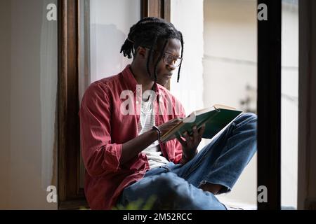 Fokussierter afrikanischer Schülerfreund mit Brille, der auf Fensterbank sitzt, ein Buch liest und sich auf die Prüfung vorbereitet Stockfoto