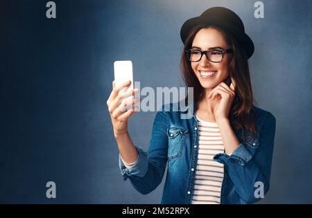 Dein bestes Lächeln macht das beste Selfie. Studioaufnahme einer schönen jungen Frau, die ein Selfie vor blauem Hintergrund macht. Stockfoto