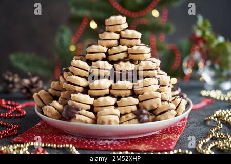 Hausgemachte Weihnachtskekse gefüllt mit Marmelade und in Schokolade getaucht, mit Tannenbaum im Hintergrund Stockfoto