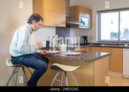 Ein älterer weißer Mann sitzt auf einem Hocker in der Küche, trinkt eine Tasse Kaffee und sieht auf das Handy. Stockfoto