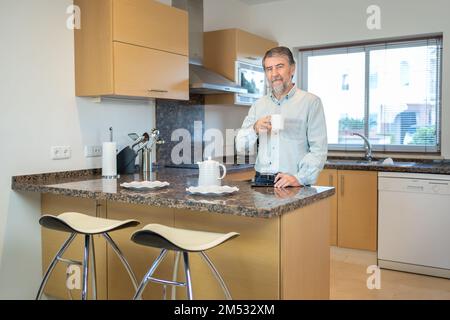 Ein älterer weißer Mann trinkt zu Hause eine Tasse Kaffee in der Küche und schaut in die Kamera. Stockfoto