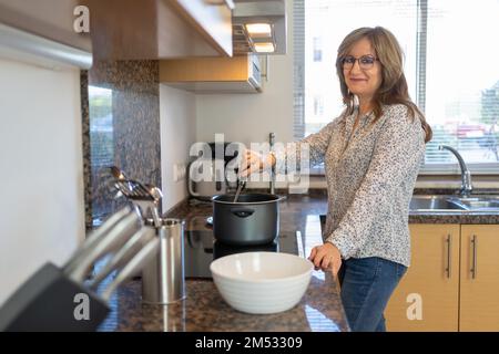 Reife weiße Frau, die zu Hause kocht und mit einer fröhlichen und lächelnden Einstellung in die Kamera schaut. Stockfoto