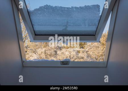 Blick vom schneebedeckten Dormerfenster auf dem Dach des Hauses bis hin zu schneebedeckten Waldbäumen an frostigen sonnigen Wintertagen. Schweden. Stockfoto