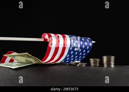 Das Konzept des Niedergangs der US-Wirtschaft. Die amerikanische Flagge liegt auf schwarzem Hintergrund neben US-Dollar und -Münzen Stockfoto