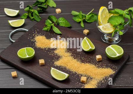 Braune Zucker- und Limettenscheiben an Bord. Minzzweige auf dem Tisch. Zitrone in Glasschüssel. Draufsicht. Dunkler Holzhintergrund. Stockfoto