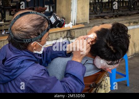 Ein Straßenfriseur in Hanoi, Vietnam, reinigt sorgfältig Ohrenschmalz aus dem Ohr eines Kunden. Stockfoto