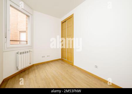 Schlafzimmer mit einem zweiteiligen eingebauten Kleiderschrank mit Eichenschiebetüren in einer Ecke und Holzfußboden und einem Fenster mit einem weißen Aluminiumheizkörper darunter Stockfoto