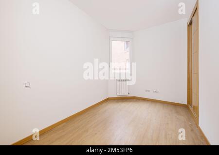 Schlafzimmer mit einem zweiteiligen eingebauten Kleiderschrank mit Eichenschiebetüren in einer Ecke und Holzfußboden und einem Fenster mit einem weißen Aluminiumheizkörper darunter Stockfoto