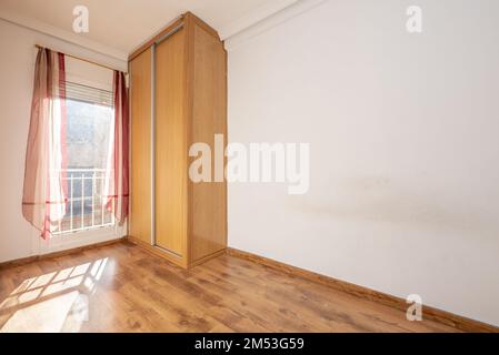 Leerer Raum mit eingebautem Holzschrank mit Schiebetüren mit Aluminiumkanten neben einem Fenster mit Vorhängen Stockfoto