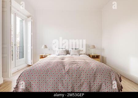 Doppelzimmer mit Queensize-Bett, Nachtschränkchen aus Holz mit zwei Lampen, Balkonfenster mit schmiedeeisernem Geländer und weißen Holzläden Stockfoto
