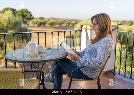 Reife weiße Frau, die an einem sonnigen Tag auf einer Apartmentterrasse ein Buch liest. Stockfoto